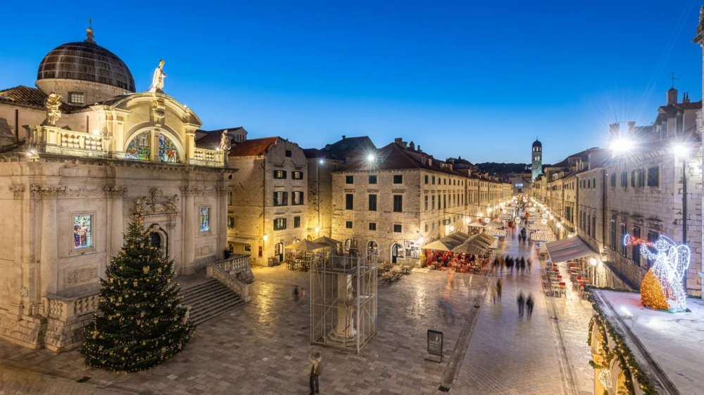 Stradun at dusk with a Christmas tree beside St Blaise's Church and festive lights along the street